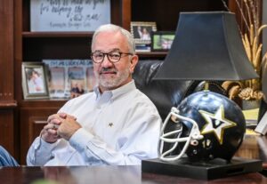 Kenny Cole sits at his office desk smiling, with his Vanderbilt football helmet — now a lamp — displayed beside him