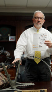 Attorney Kenny Cole stands in his office holding a tree stand's printed assembly instructions, with the product's camouflage and metal frame visible in the foreground